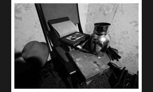Een monochrome foto toont een intieme hoek van Villa Hector, een verlaten huis in België. Twee stoelen staan naast elkaar; één heeft afgeronde contouren, terwijl de andere een rechte rugleuning heeft met een open boek erop. Een decoratieve metalen urn rust op een patroonrijke doos of kleine koffer naast deze stoelen, allemaal tegen een vintage bloemenbehang dat spreekt van het vroegere grandeur van het huis.